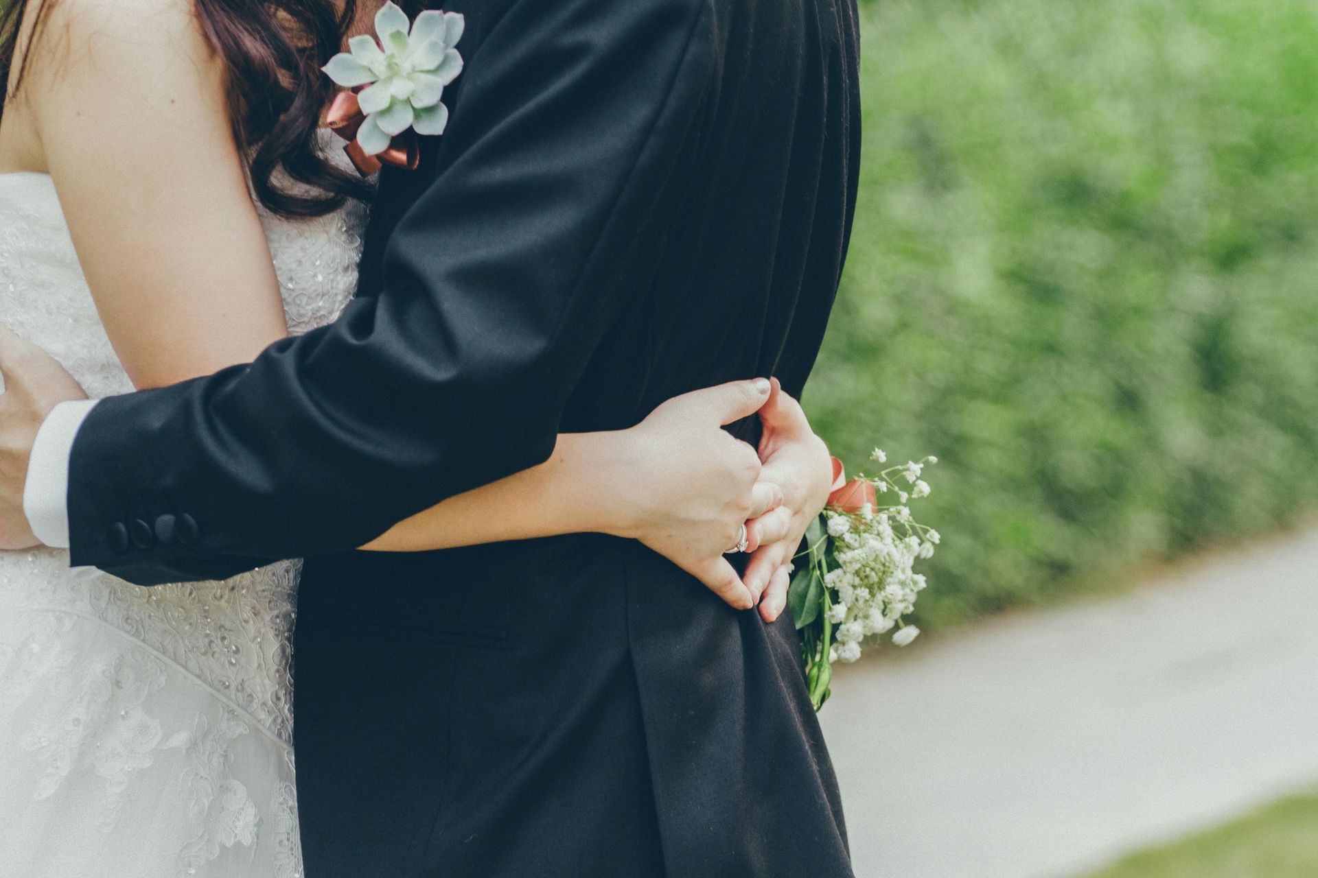 Couple in wedding attire hugging outdoors. Bride in white dress, groom in black suit.