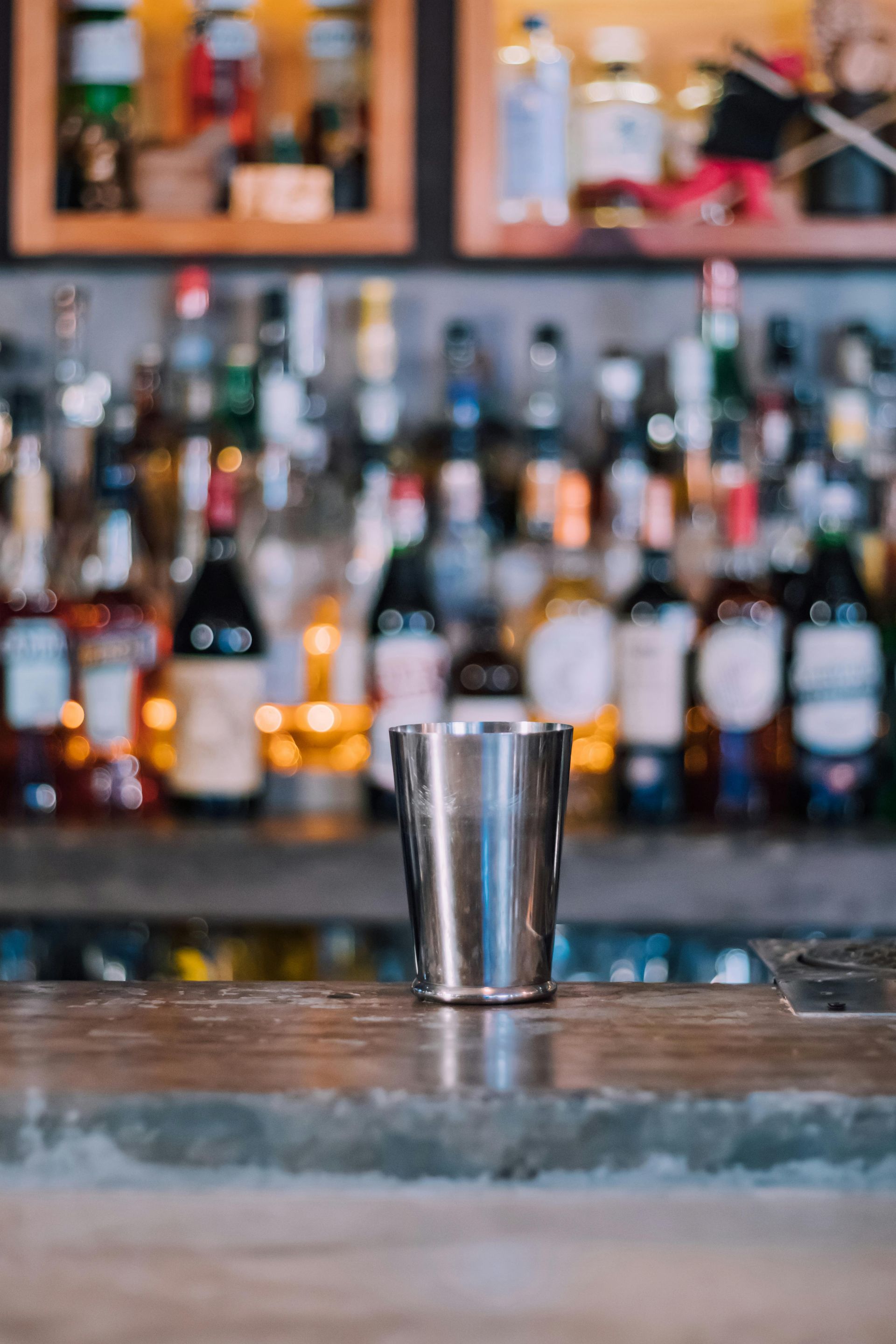 Stainless steel cocktail shaker on a bar, bottles blurred in background.