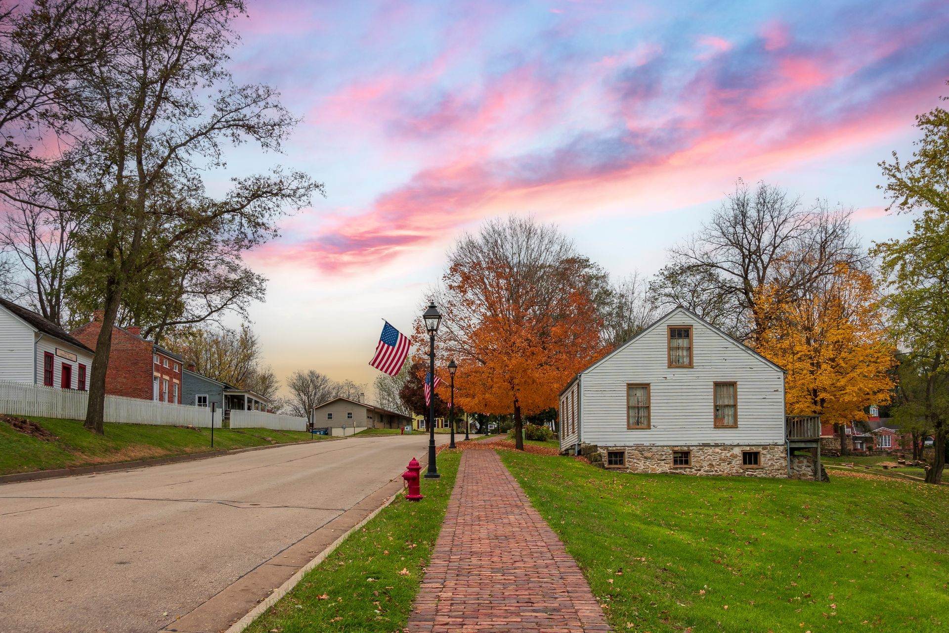 Brick sidewalk leading past a white house and autumn trees under a pink sunset sky.