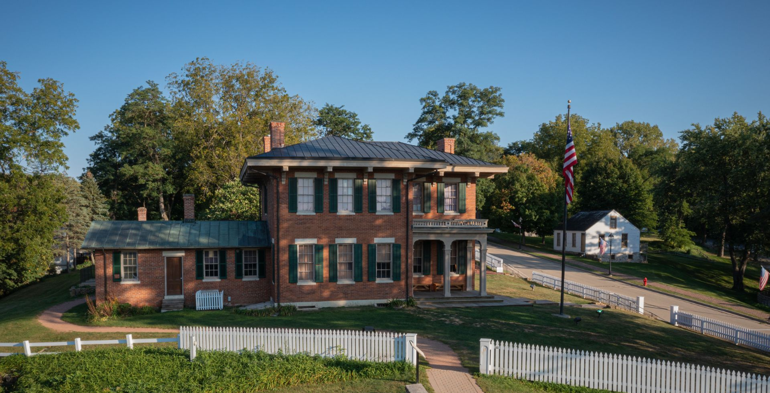 Historic brick house with green roof on a sunny hillside, surrounded by trees and white fencing.