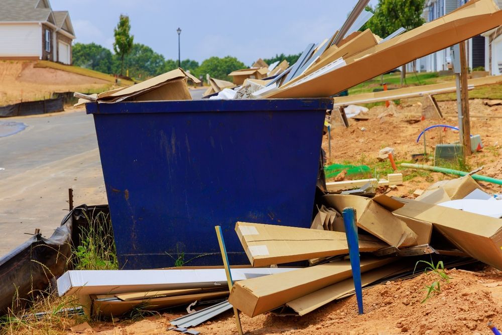 A blue construction dumpster overflowing with cardboard and debris on a dirt lot near new housing.
