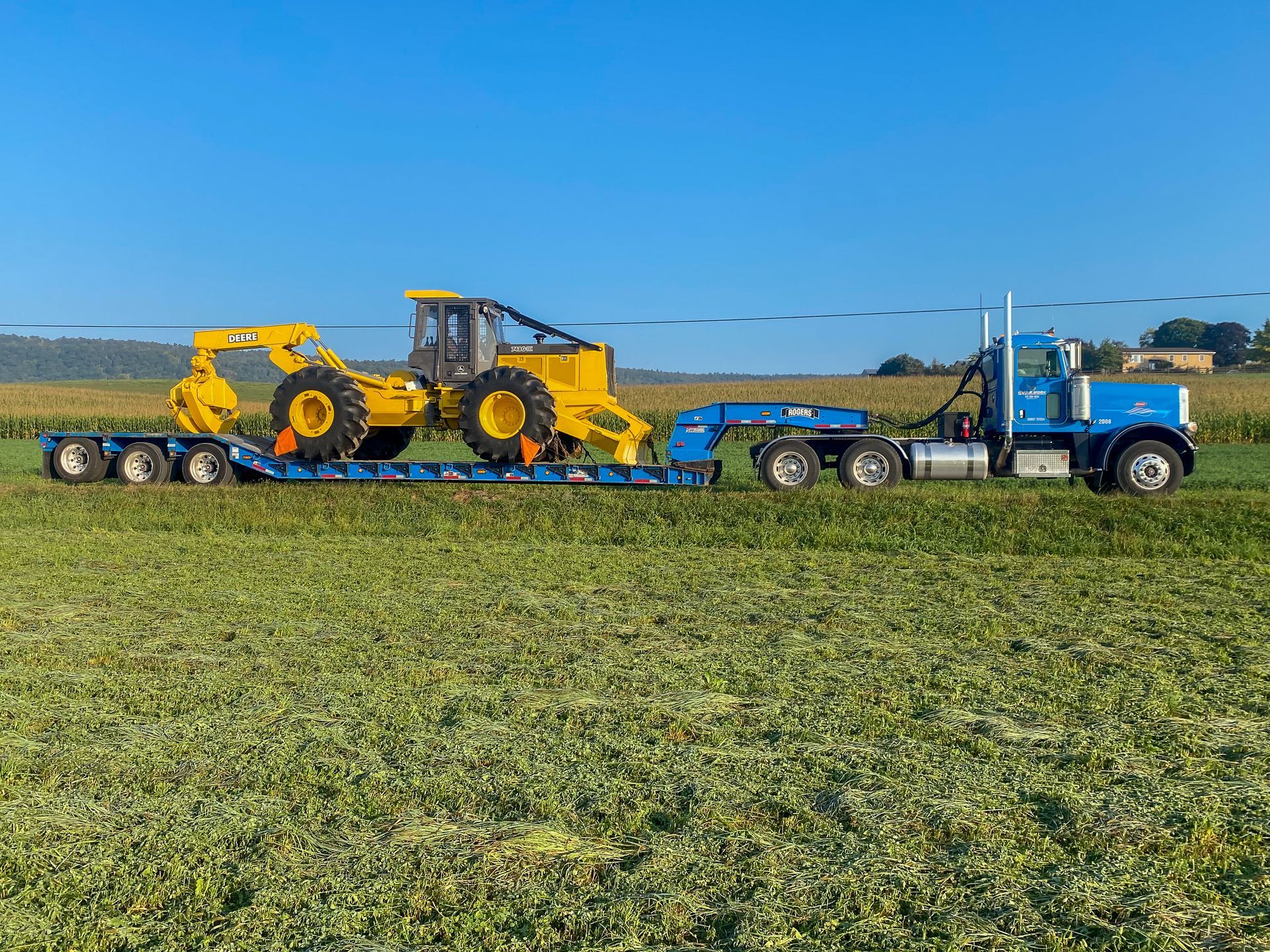 A blue semi truck from R. Work Excavating & Trucking is carrying a yellow tractor on a trailer in a field.