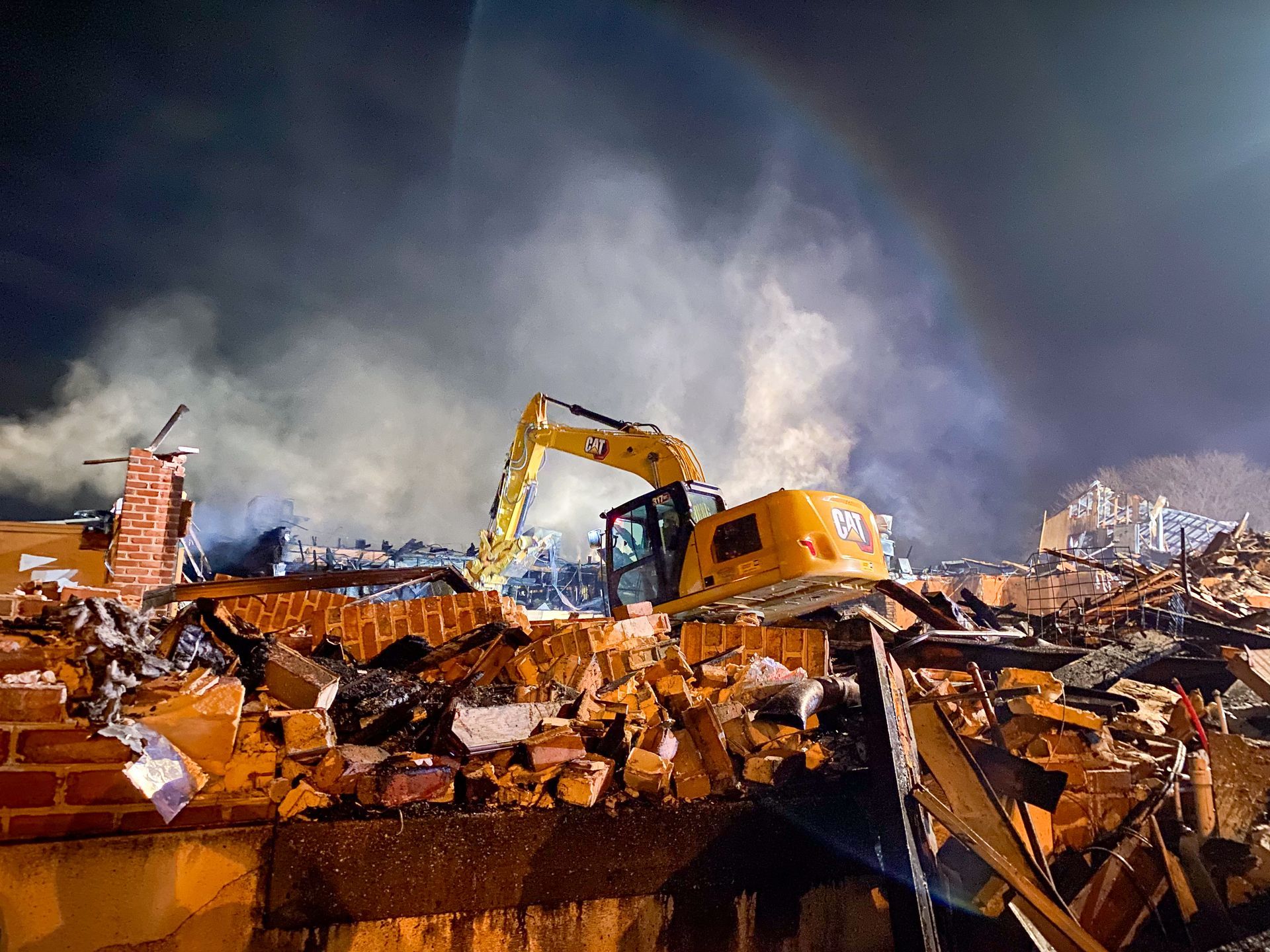 A yellow excavator from R. Work Excavating & Trucking is demolishing a building with smoke coming out of it.