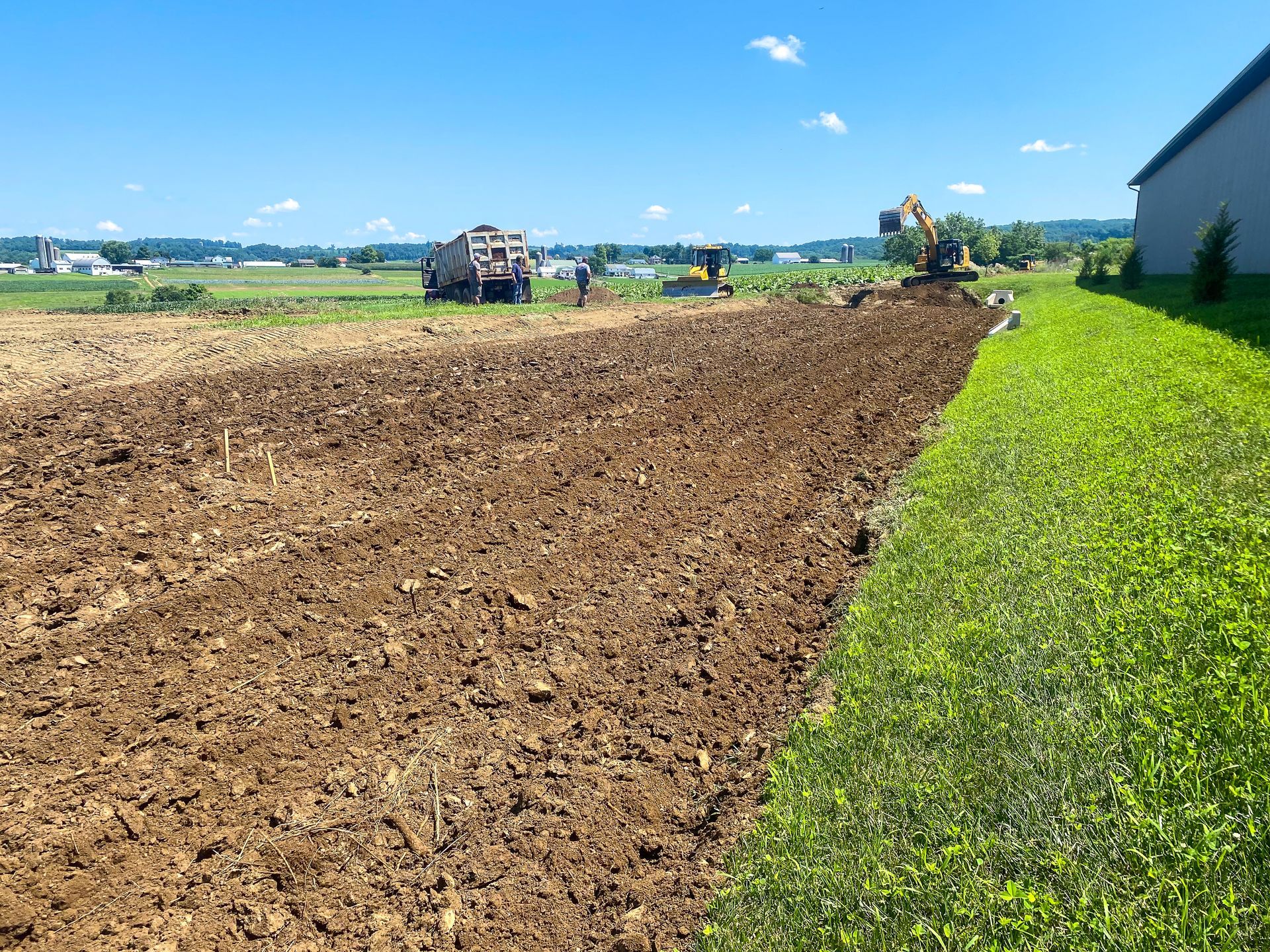 A dump truck and two excavators from R. Work Excavating & Trucking are plowing a field next to a building.