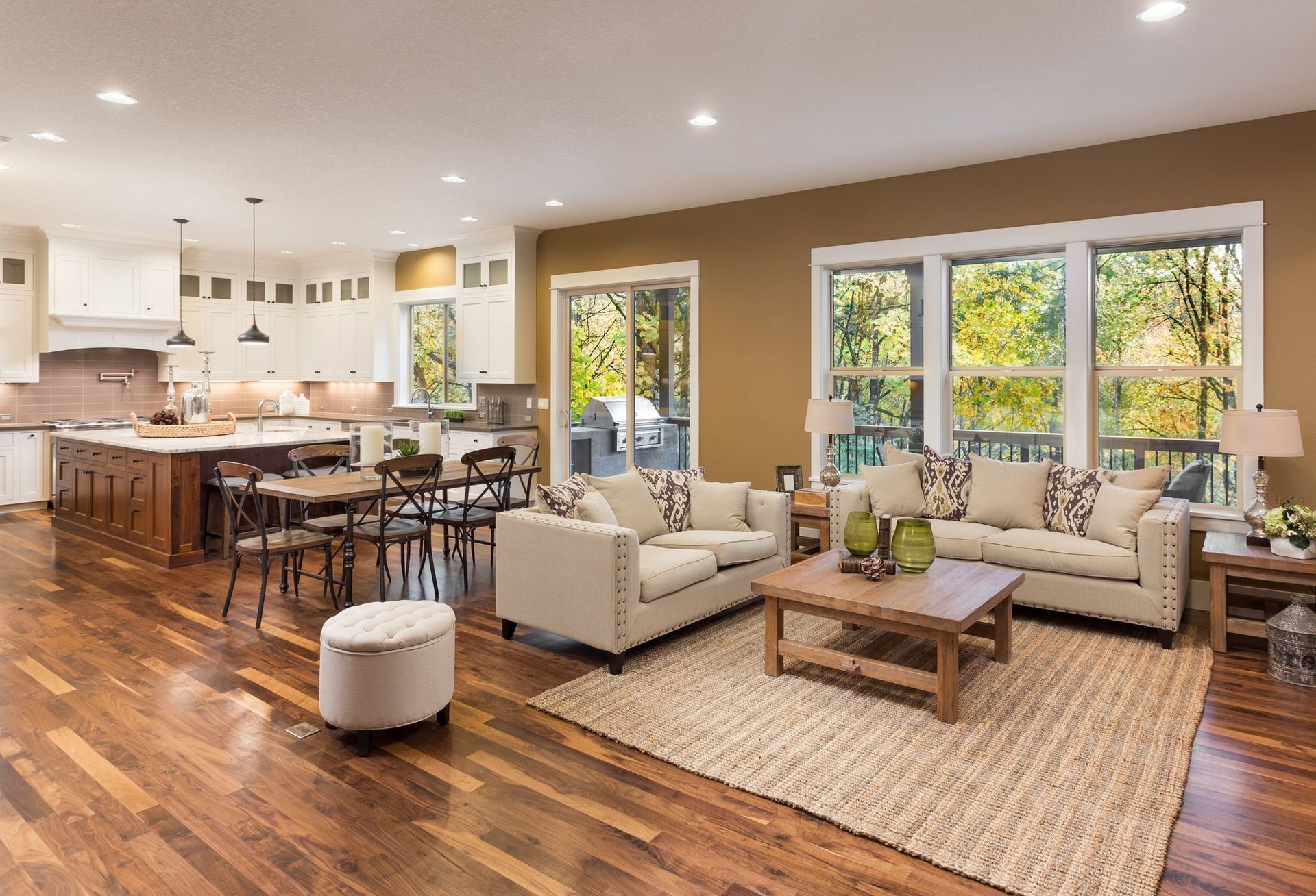 Open-plan living space with kitchen, dining, and living areas. Brown wood floors, beige sofas, and a view of trees.