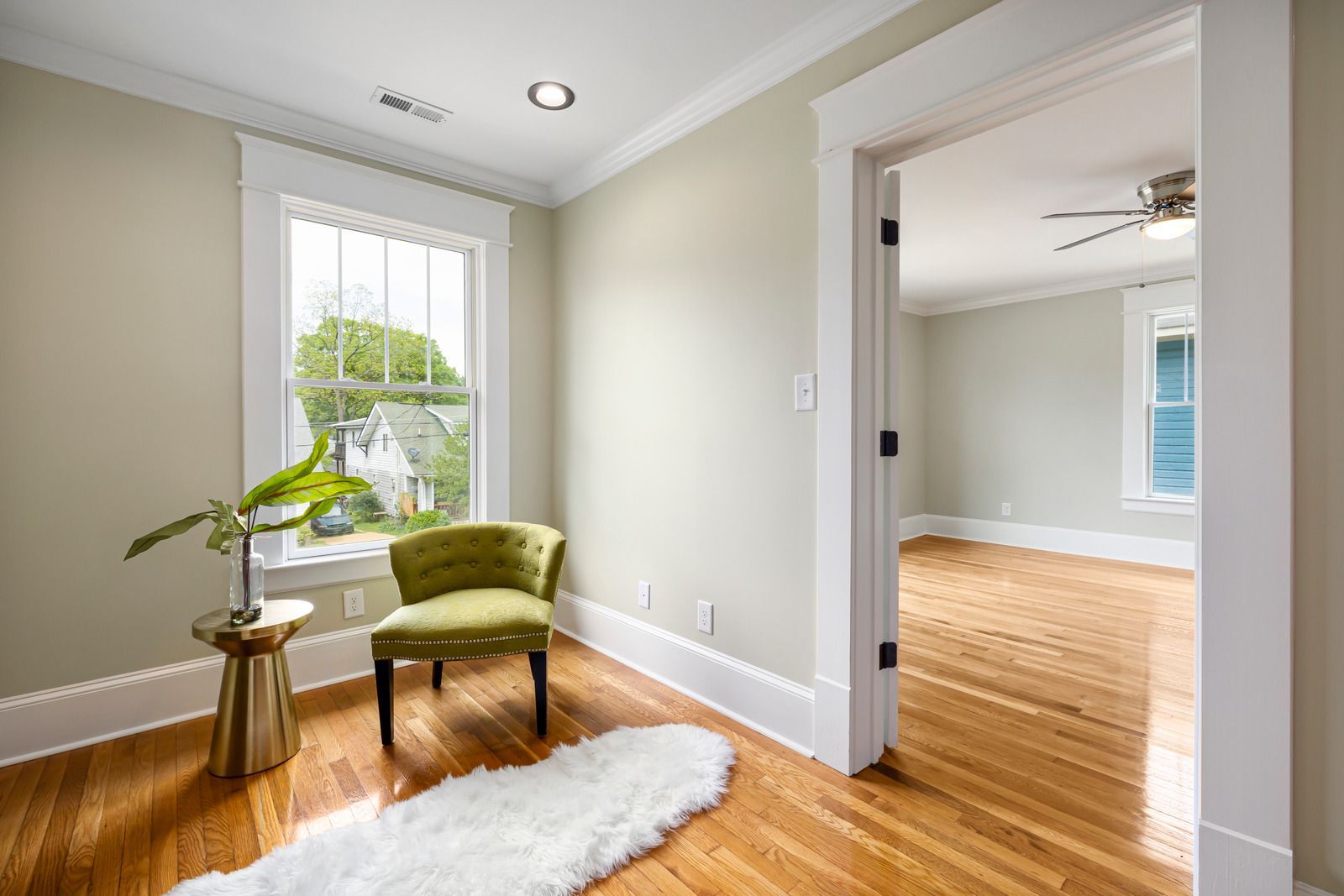 Bright empty room with green chair, potted plant, hardwood floors, and adjoining room visible through wide doorway