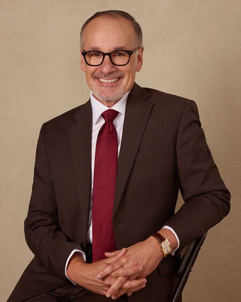 Smiling person in a brown suit with red tie, seated against a plain beige background.
