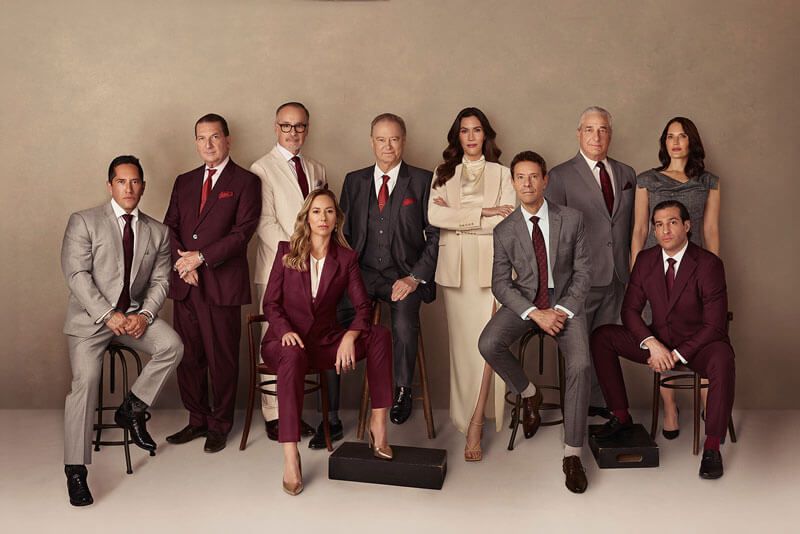 Group portrait of ten people in formal attire posing in a studio against a beige backdrop