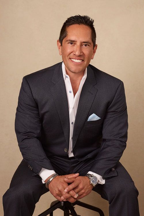 Man in a dark suit and white shirt seated against a beige backdrop, smiling at the camera.