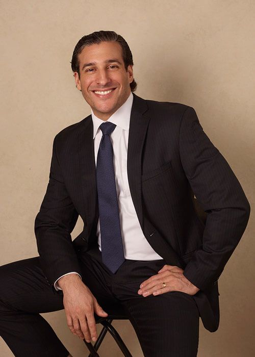 Smiling person in a dark suit and tie seated against a beige background.