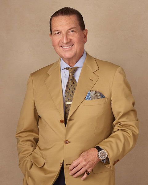 Man in a tan suit smiling, posing against a plain beige background.