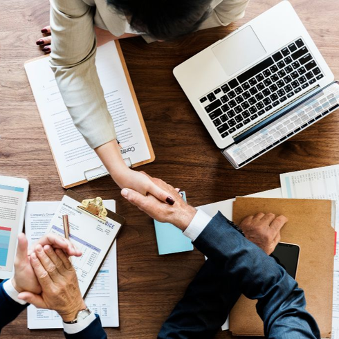 Two people shaking hands at a desk with a laptop, documents, and a clipboard.