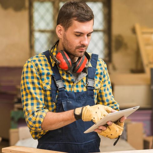 Man in a plaid shirt and apron using a tablet in a workshop with ear protection around his neck