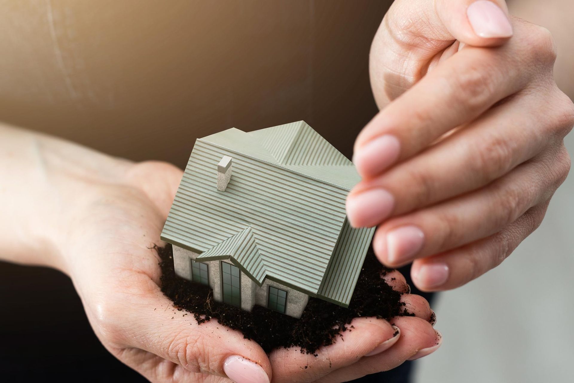Hands holding a small house model with soil, symbolizing home ownership or real estate.