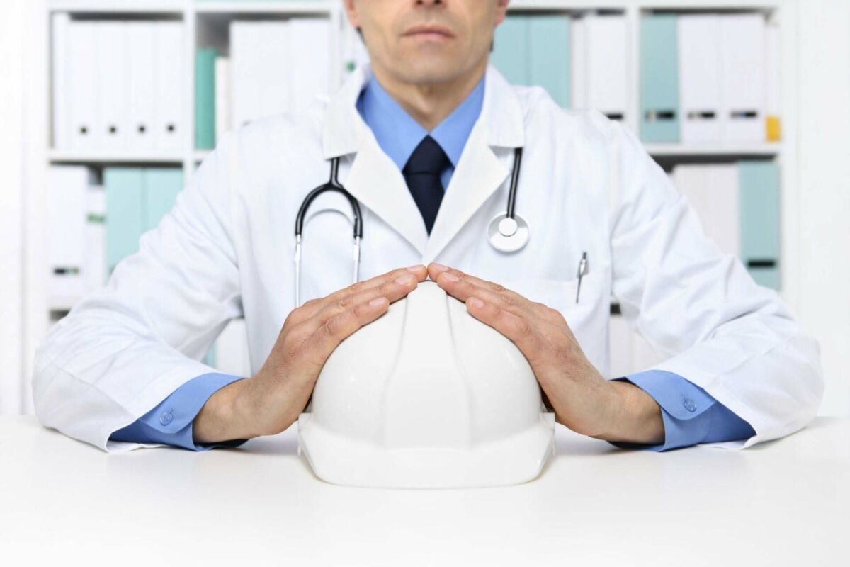 Doctor in white coat holding a white hard hat at a desk in an office setting