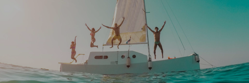 People on a sailboat deck jumping and posing above turquoise water under tall sails