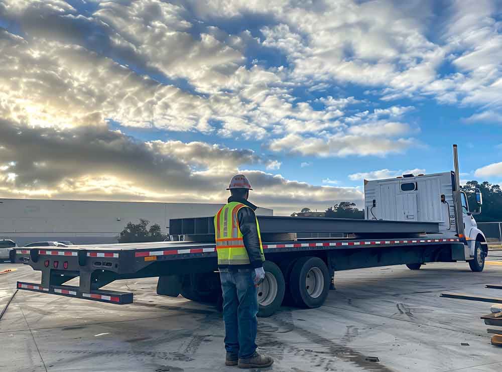 Worker in a safety vest standing beside a flatbed truck at sunrise on an industrial lot.