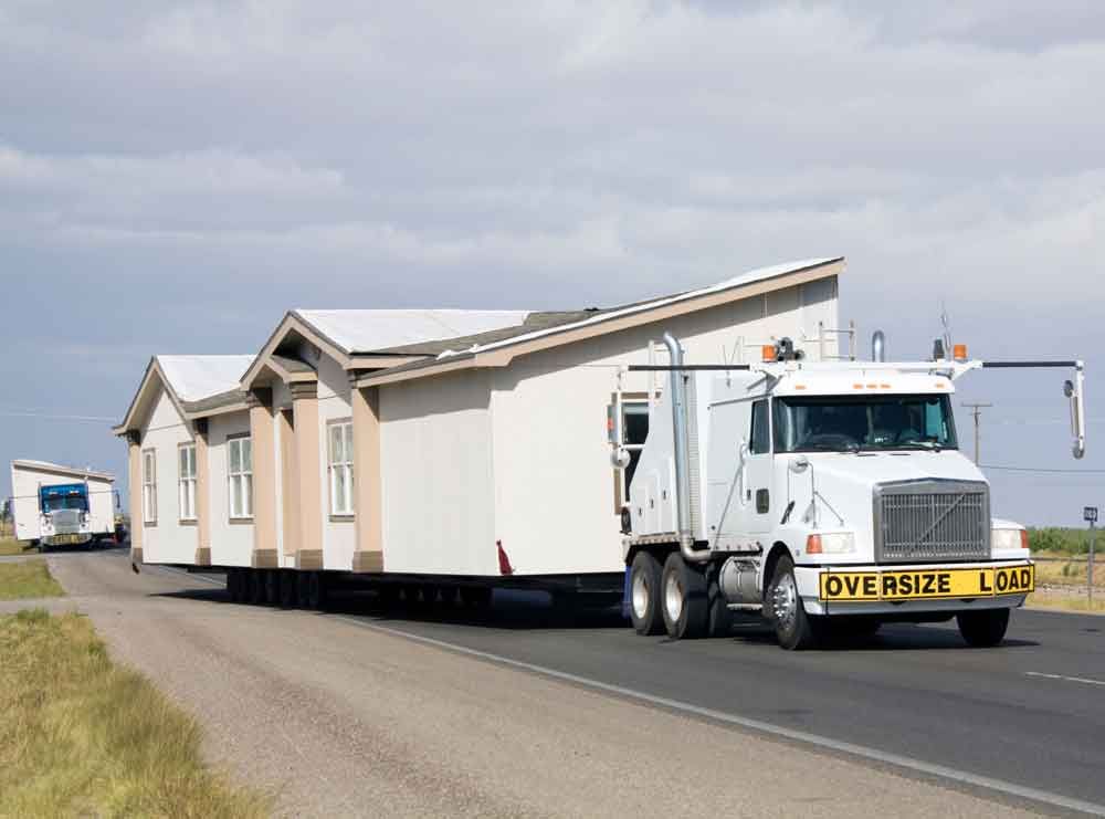 Oversize load house on a semi-truck driving along a rural road under cloudy skies