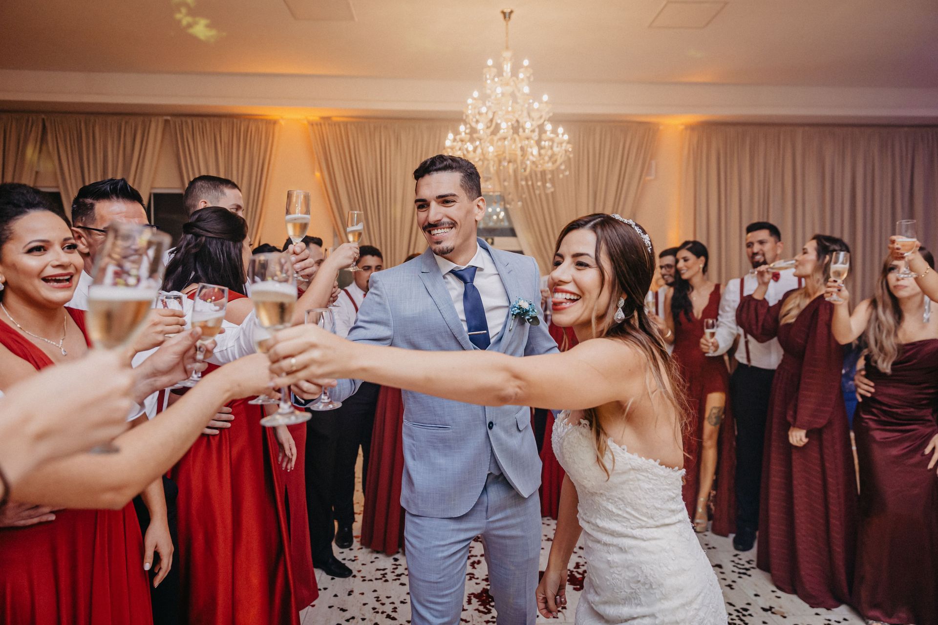 Guests in formal wear clink glasses at a wedding reception with warm lights and a chandelier.