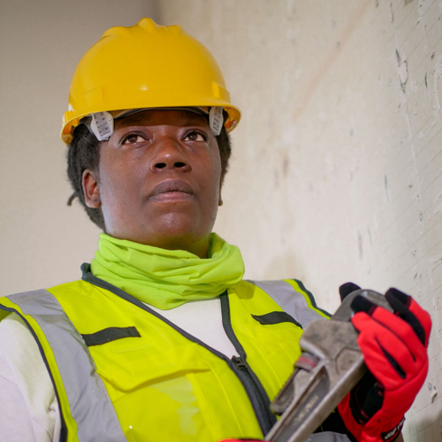 Construction worker in a yellow hard hat and vest holding a wrench indoors