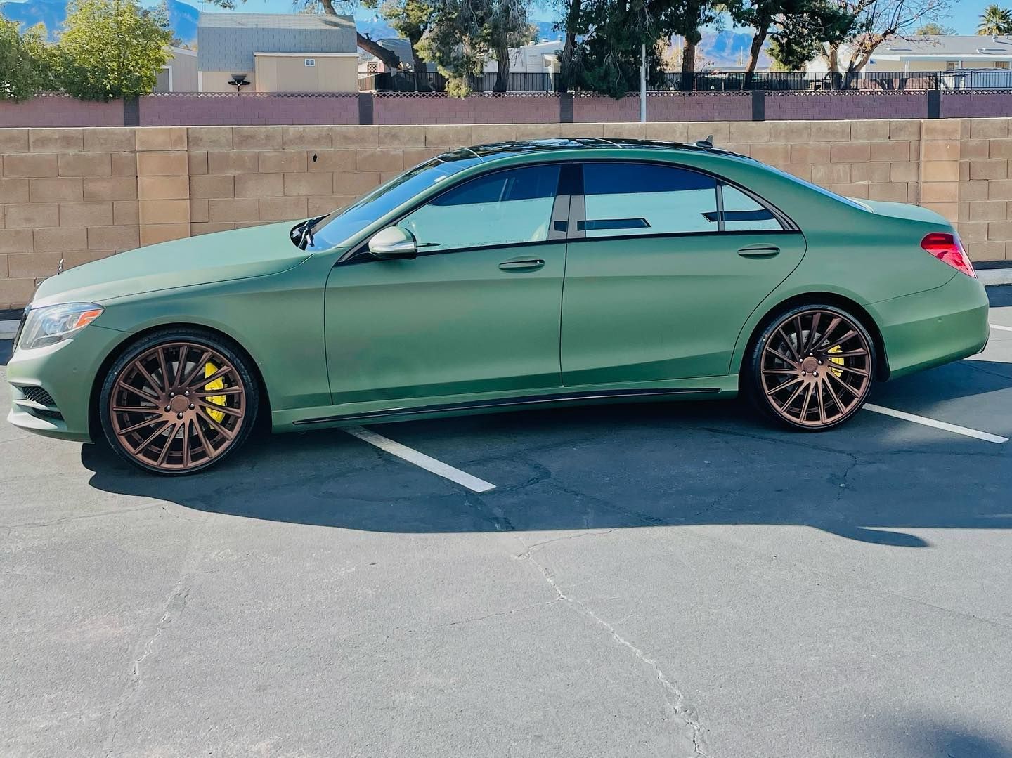 Green sedan parked in a lot, side view with black wheels and a tan brick wall behind it