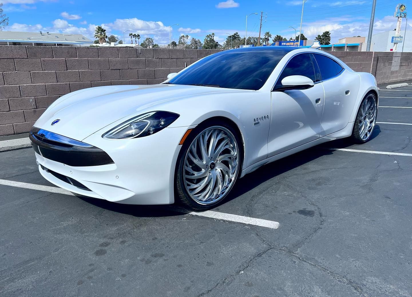 White sports car parked on asphalt beside a brick wall under a blue sky