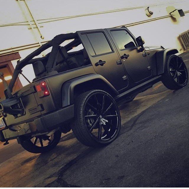 Matte black Jeep-style SUV with a roof rack and large custom wheels in a garage