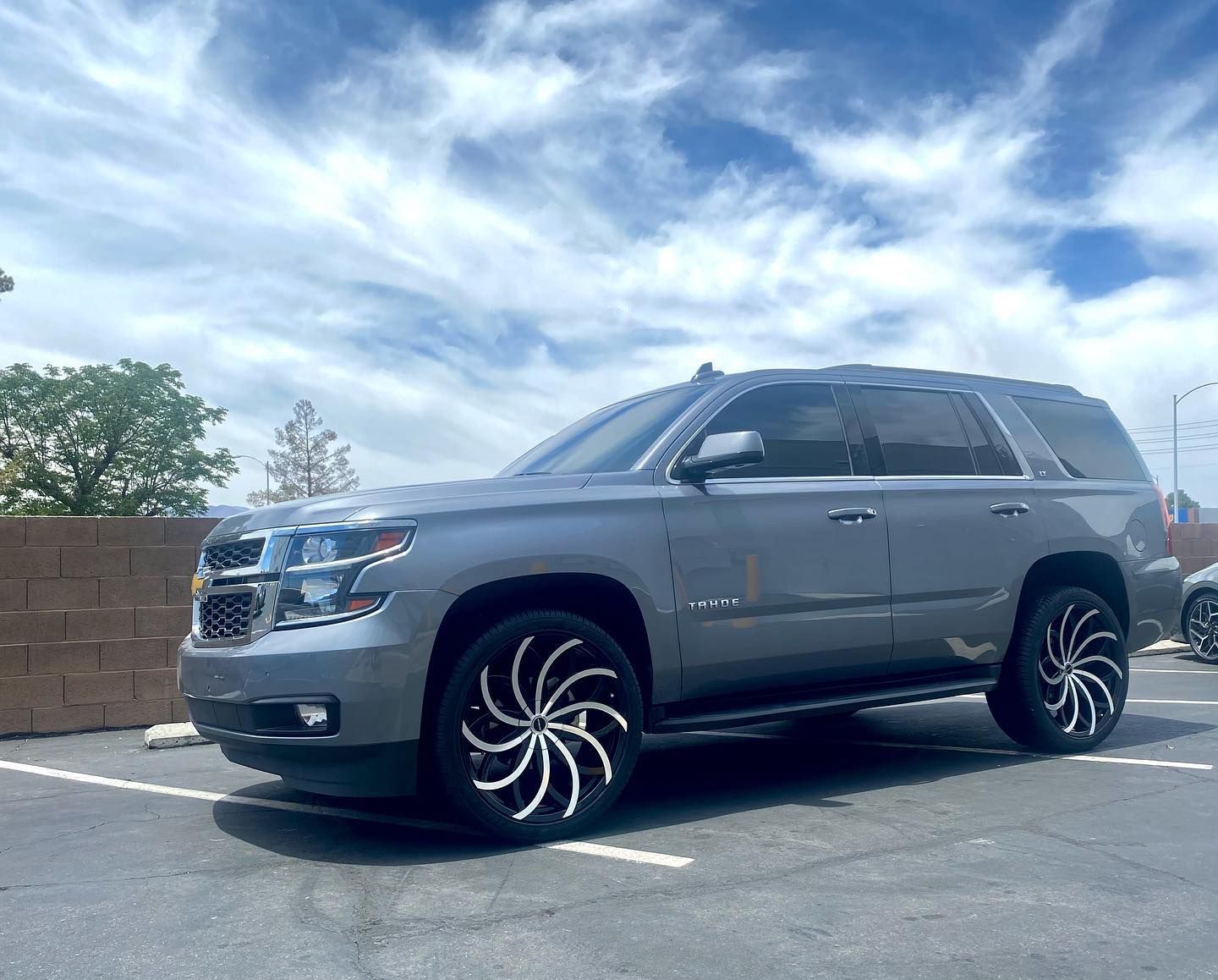 Gray SUV parked in a lot under a cloudy blue sky, with custom black swirl rims.