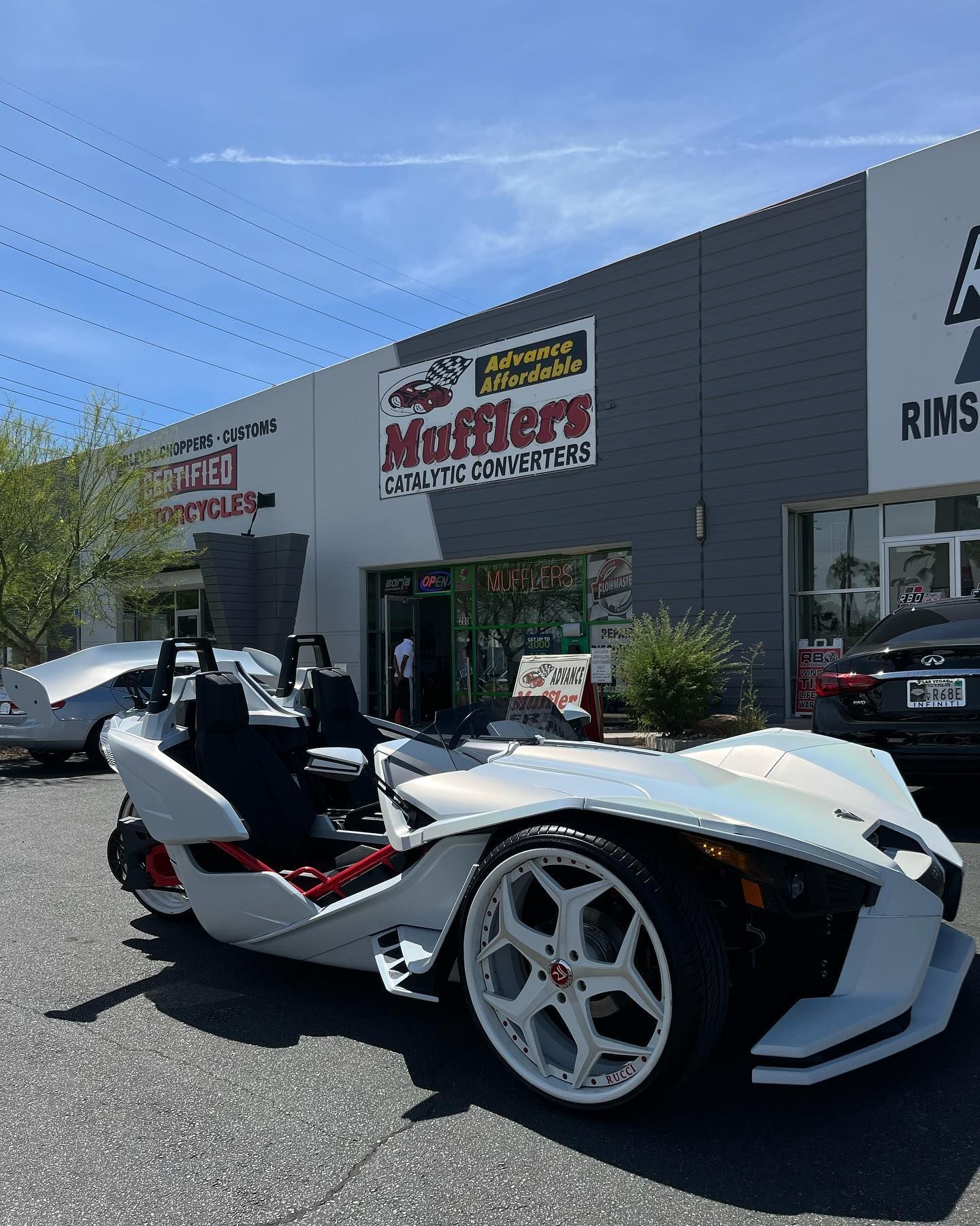 White Polaris Slingshot parked outside a motorcycle shop on a sunny day, with large white wheels.