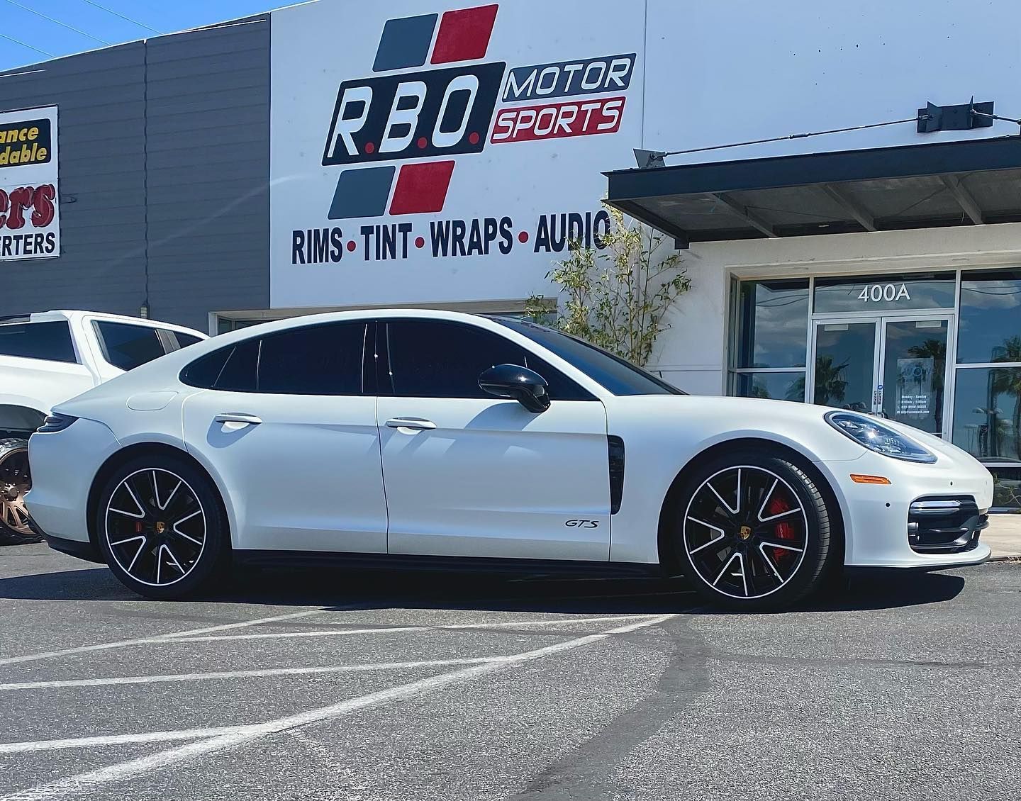 White sports sedan parked outside RBO Motor Sports storefront on a sunny day