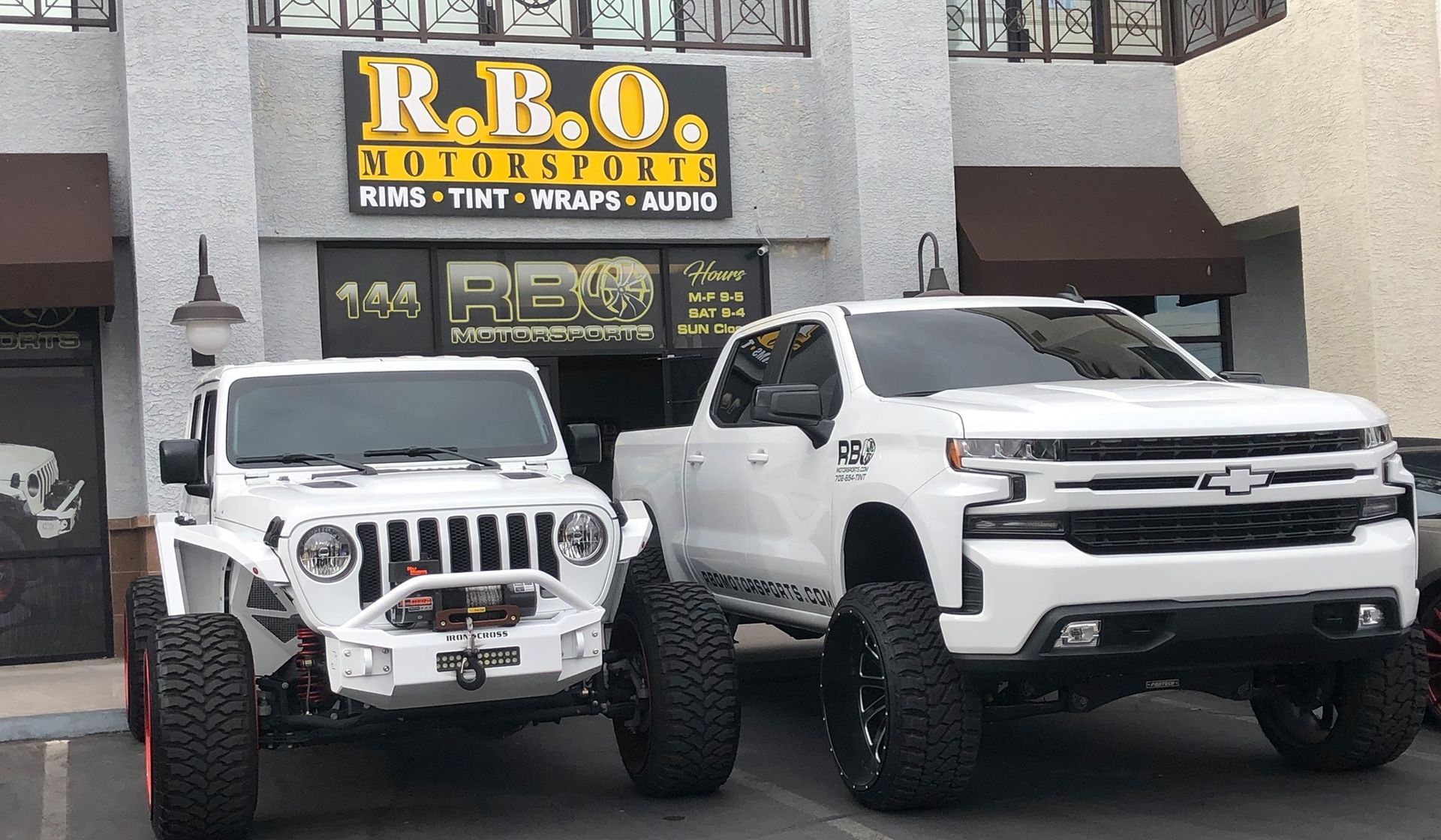 Two white lifted Jeep trucks parked outside an auto customization shop.
