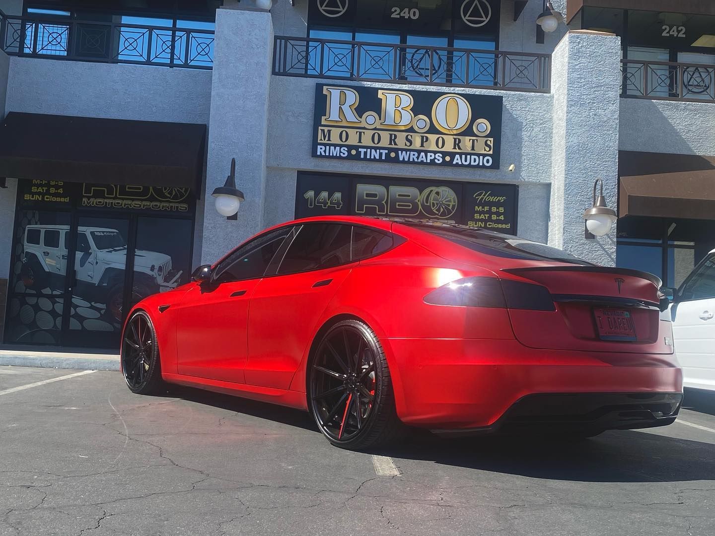 Red Tesla sedan parked outside a storefront with a black-and-gold sign.
