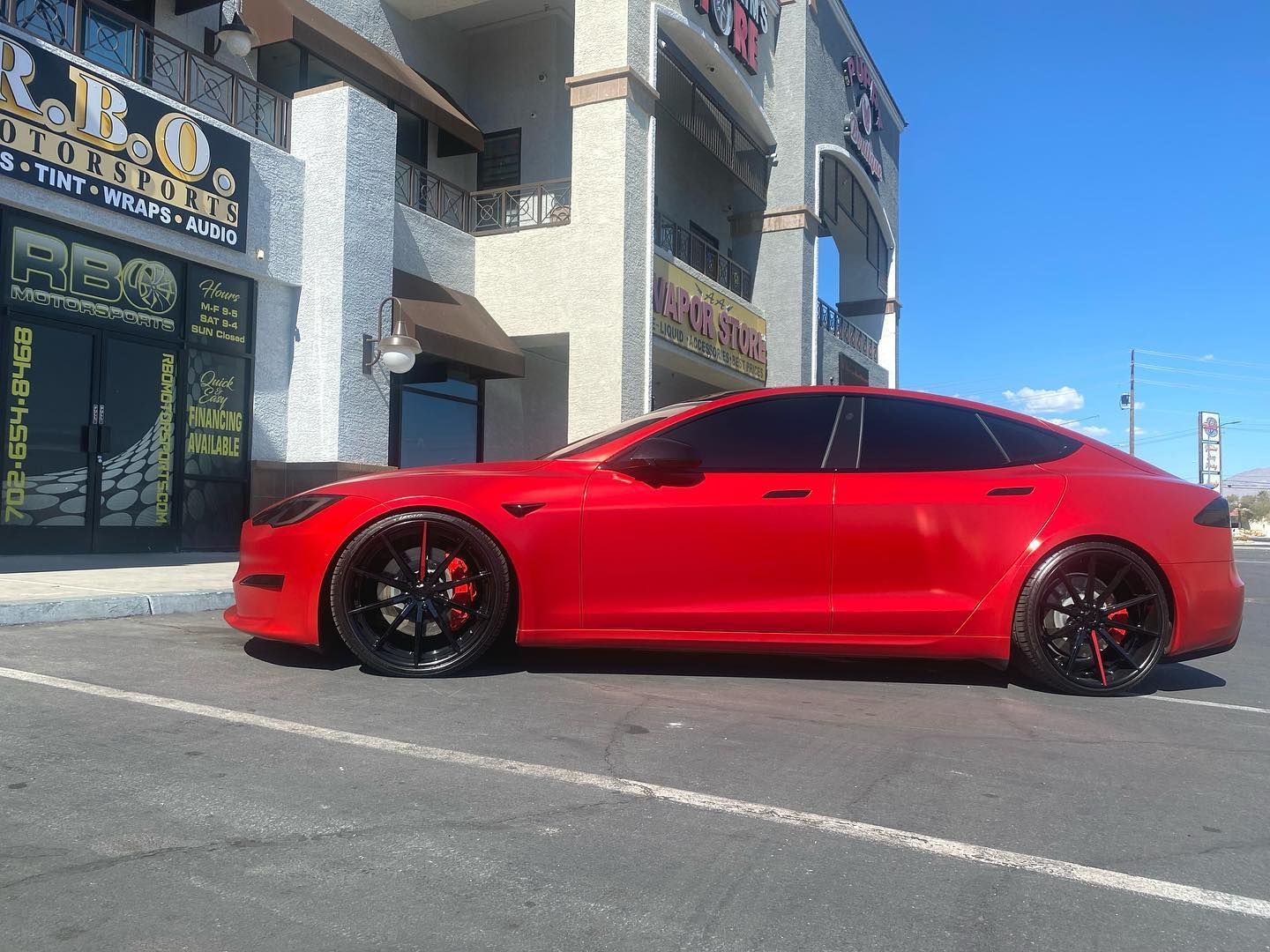 Red Tesla sedan parked beside a building on a sunny street.