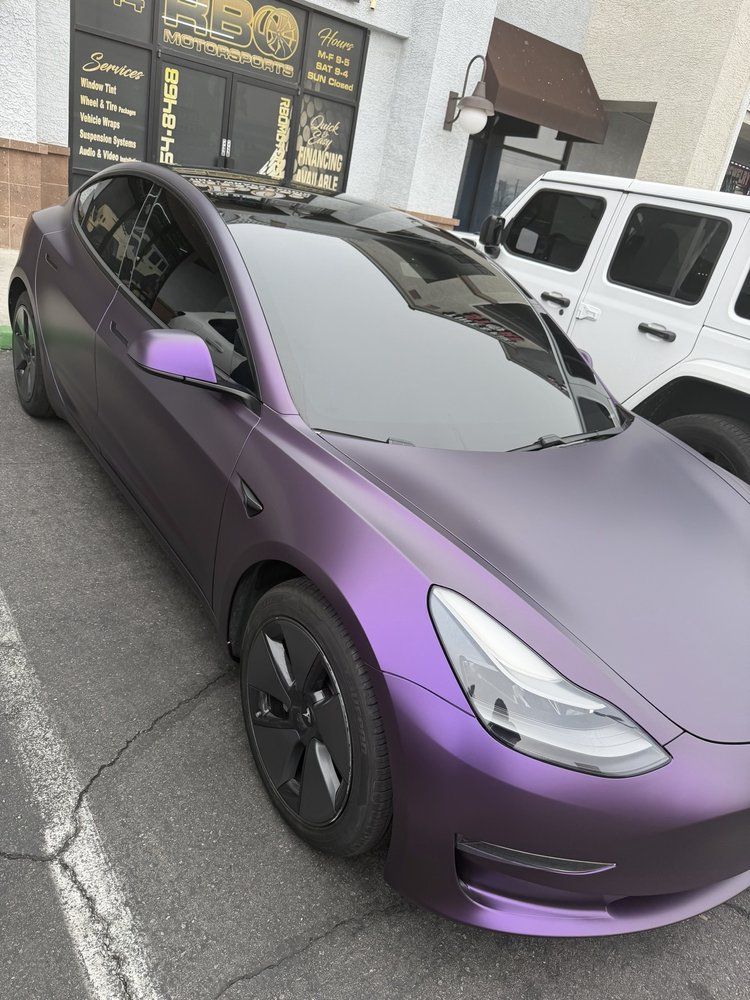 Purple matte Tesla parked on a street beside a white SUV, viewed from the front-left corner.