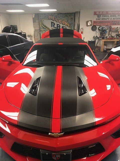 Red sports car with black and red racing stripes inside a garage showroom, viewed from the front.