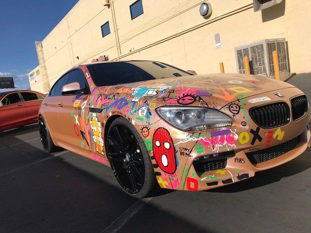 Graffiti-covered tan BMW parked outside a beige building, with black rims and a red car beside it