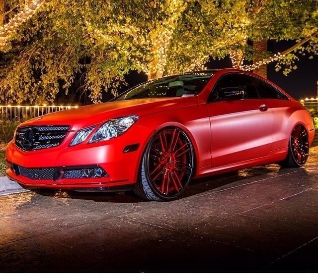 Red sports coupe parked under autumn trees at night with lights reflecting on the glossy body.
