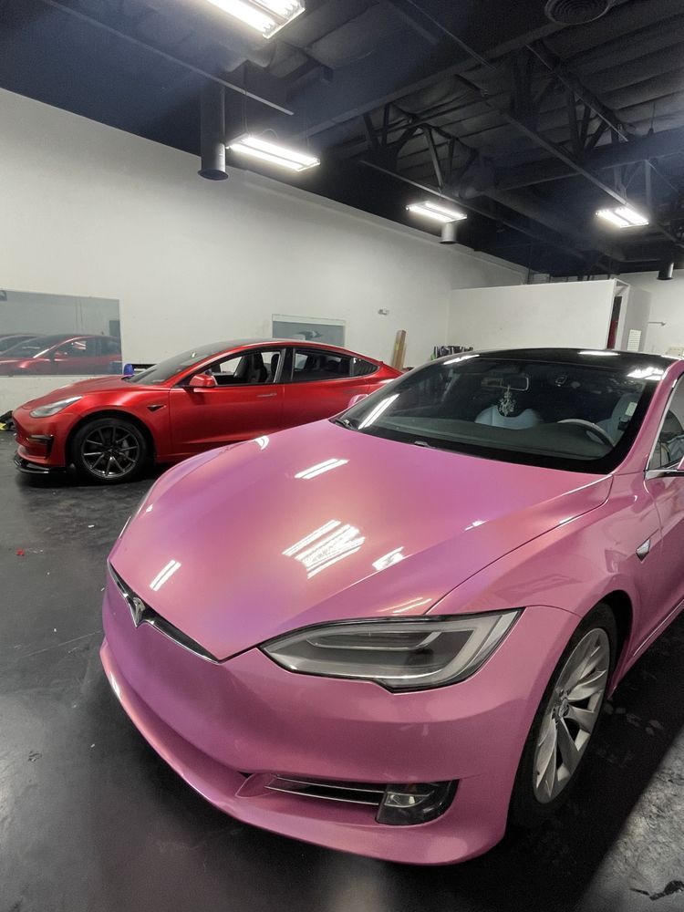 Pink Tesla sedan in a showroom with a red Tesla behind it under bright ceiling lights