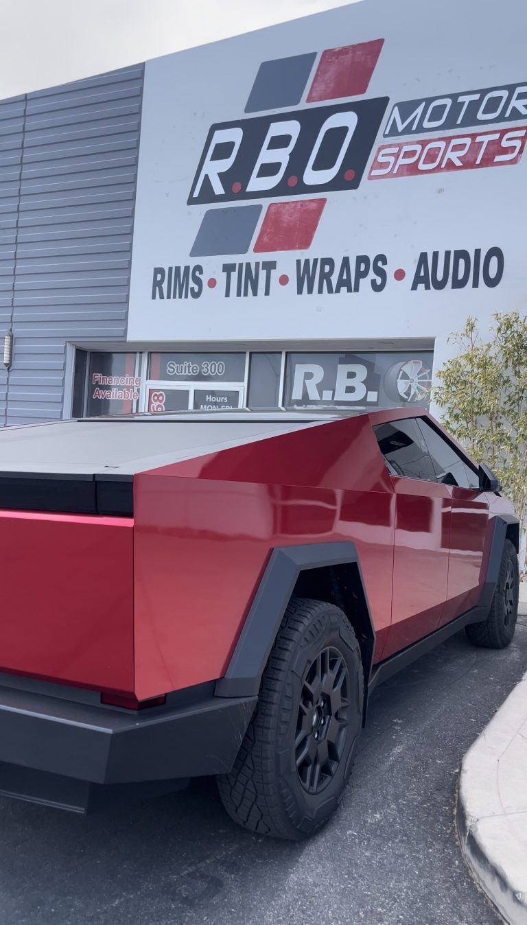 Red pickup truck parked outside RBO Motorsports shop with signage for wraps, tint, and audio.