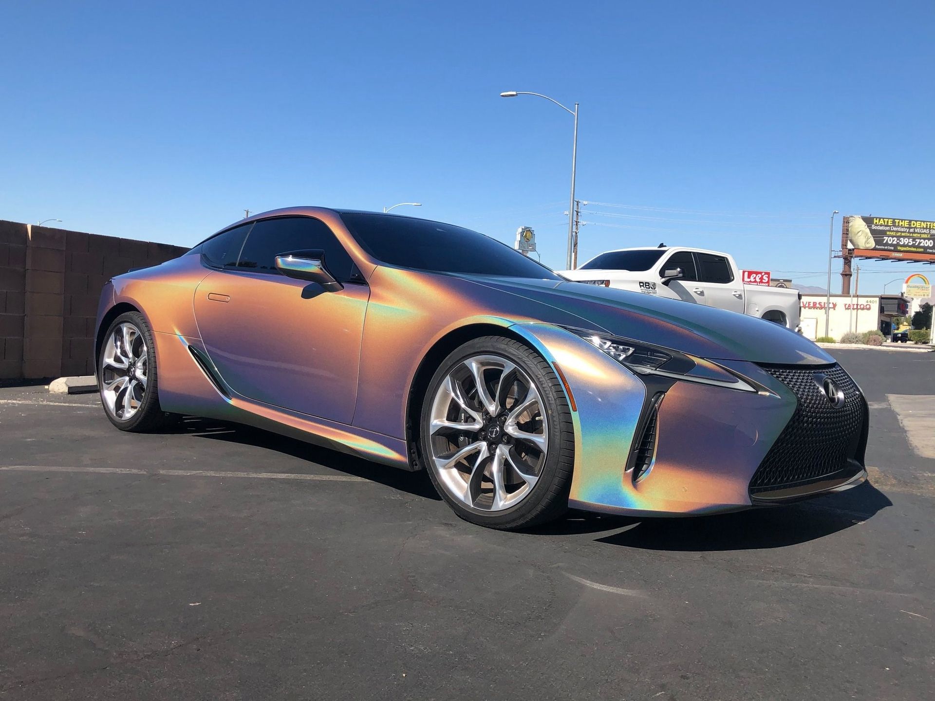 Iridescent sports coupe parked outdoors under a blue sky, viewed from the front-left.