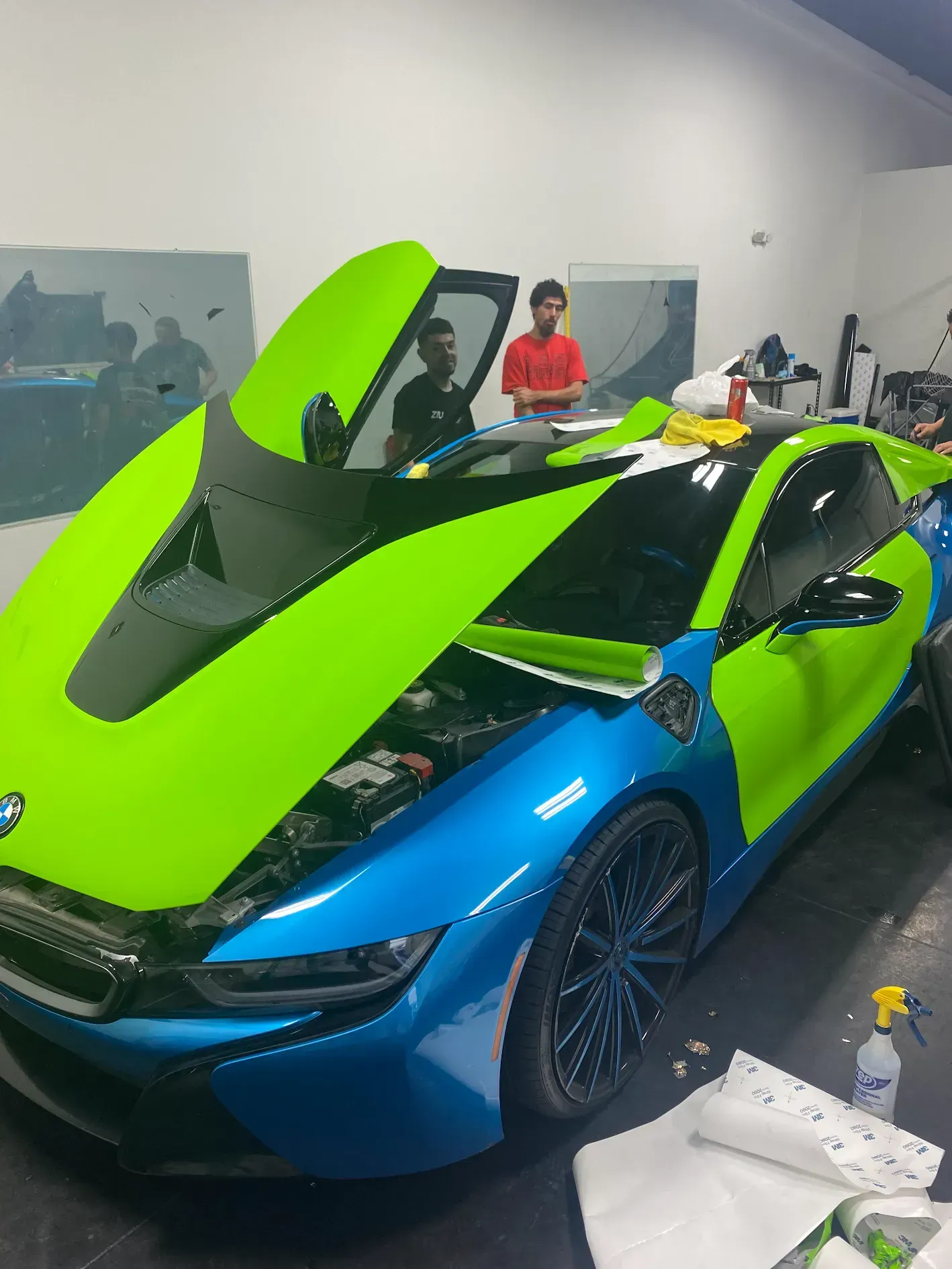 Blue-and-neon-green sports car with its hood open in a workshop, with people and tools in the background