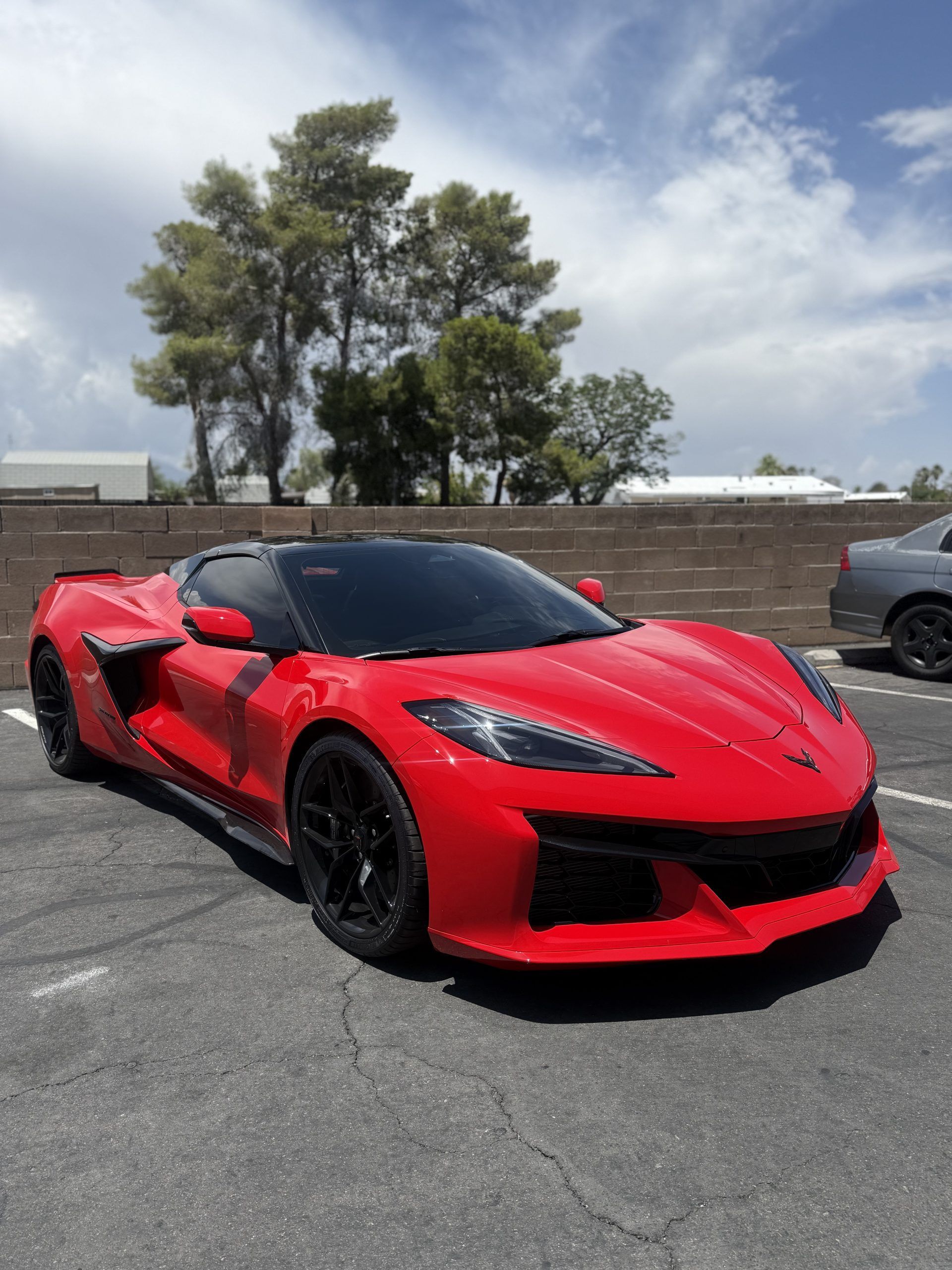 Red sports car parked on pavement under a cloudy sky, with trees and a low wall in the background.