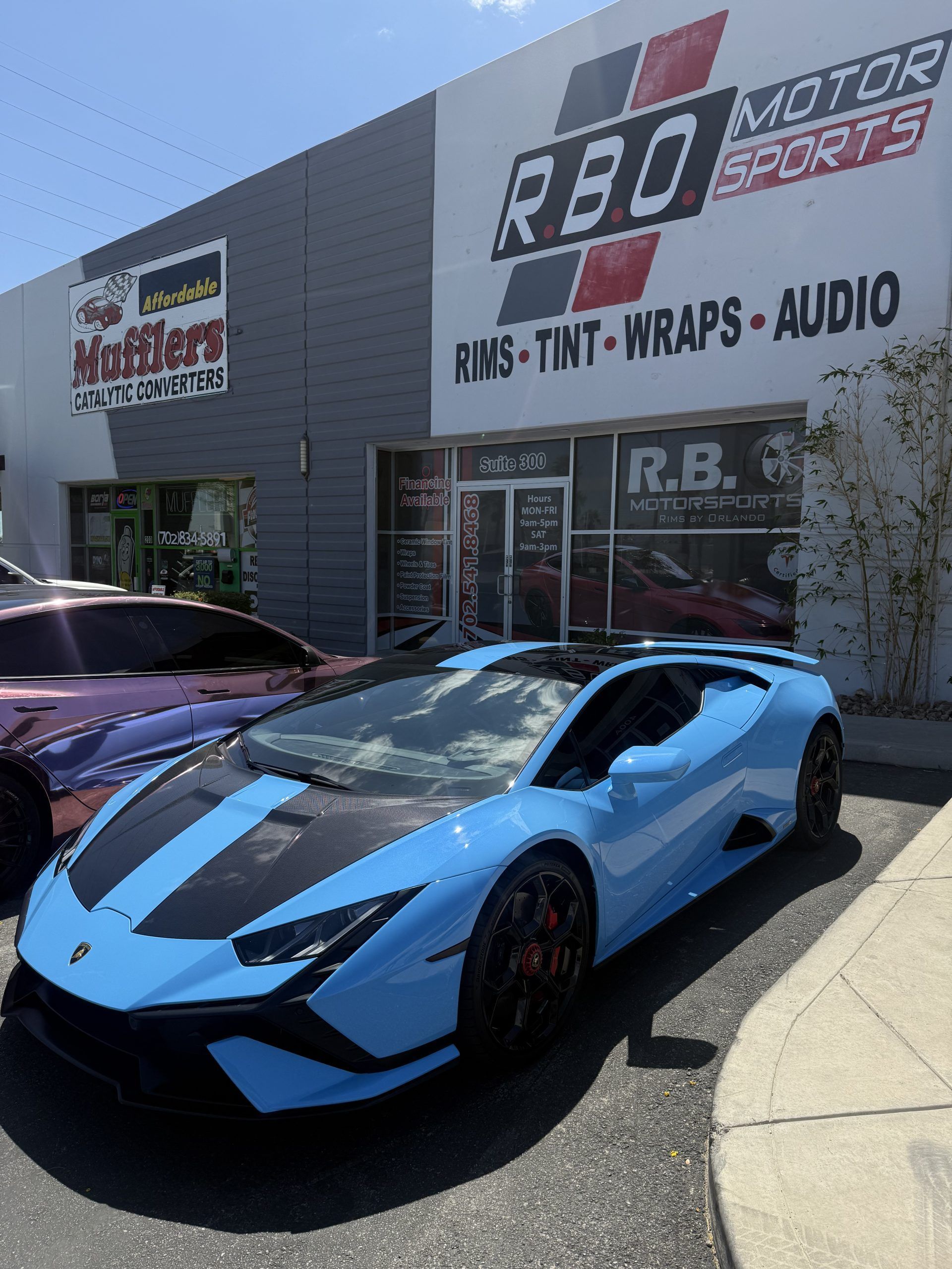 Blue sports car parked outside R&B Motor Sports shop under bright sunlight