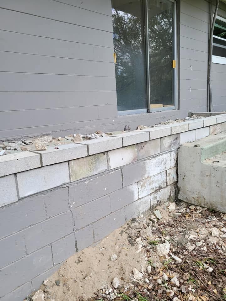 Damaged cinder block retaining wall beside a gray house, with loose debris and exposed soil.