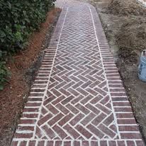 Brick walkway with herringbone center and red brick borders alongside a dirt path outdoors
