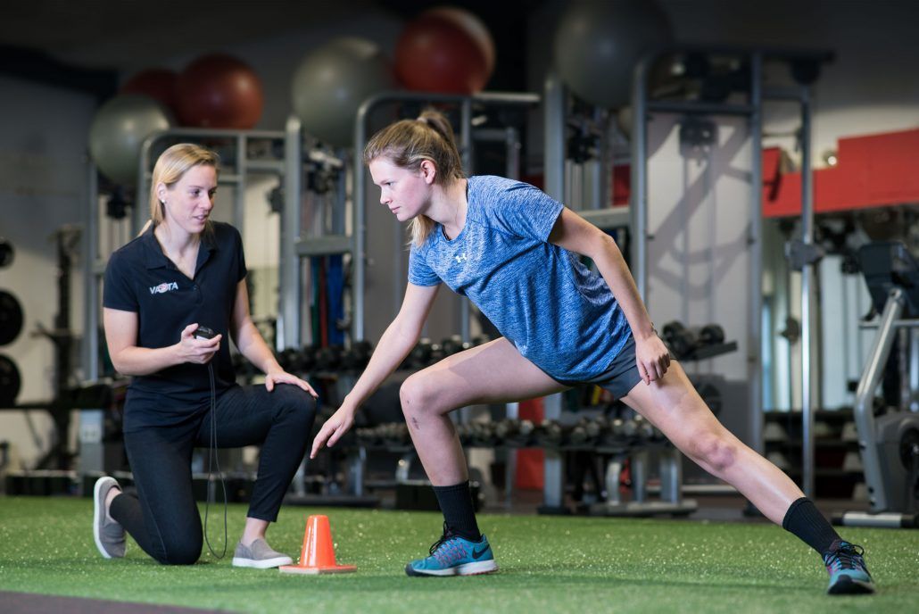 A woman is helping another woman stretch her legs in a gym.