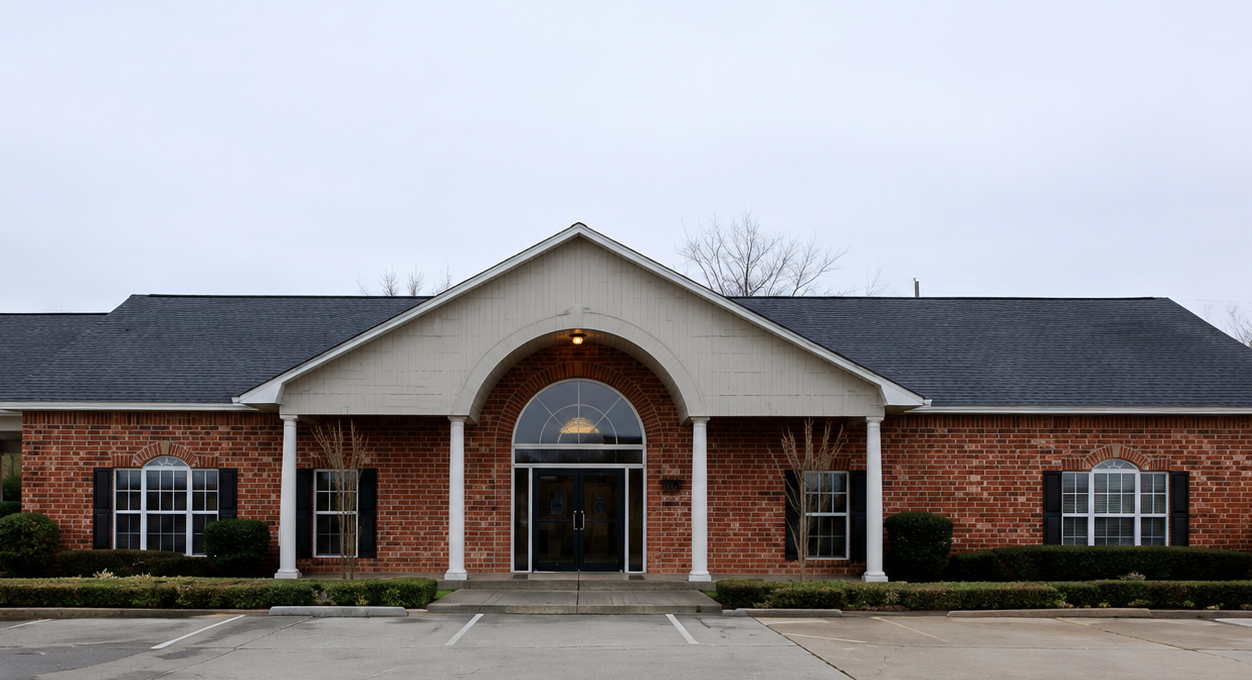 Brick building with a central arched entrance and dark roof, viewed from the front under an overcast sky