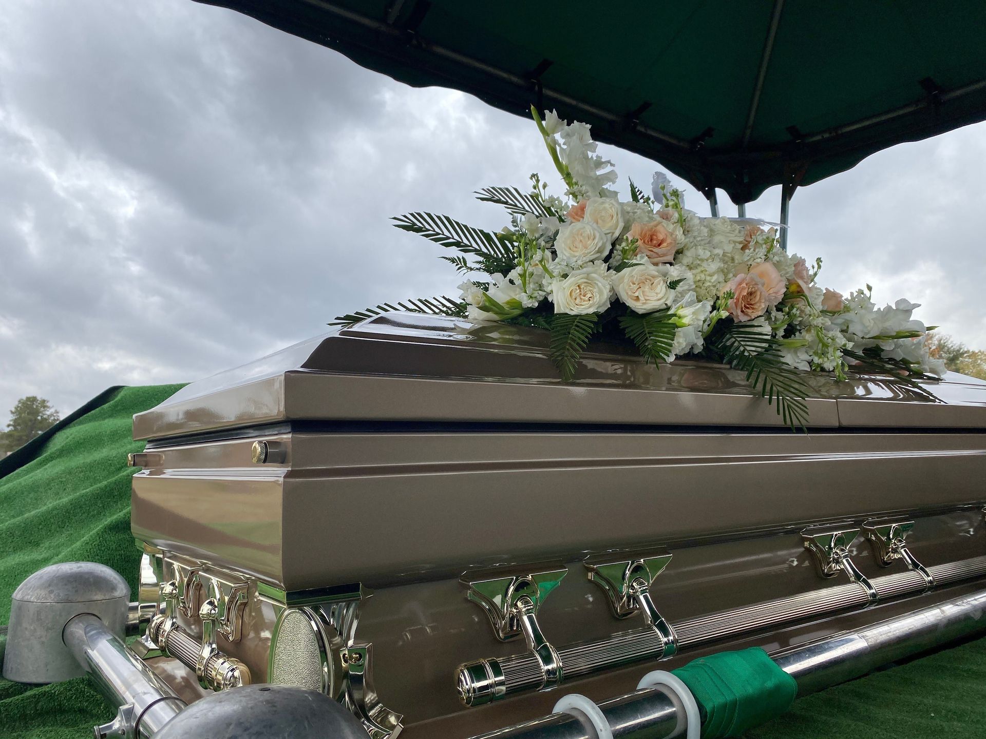 Silver casket with white and peach flowers at a graveside ceremony under a cloudy sky