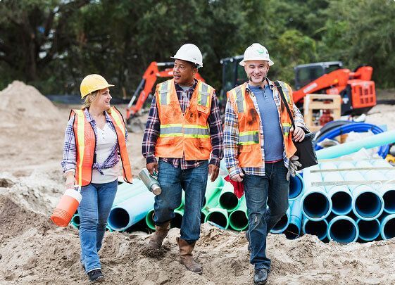 Construction workers in hard hats and safety vests standing by stacked blue pipes at a worksite