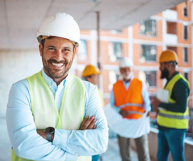Smiling construction supervisor in white hard hat and reflective vest, with workers blurred on a job site.